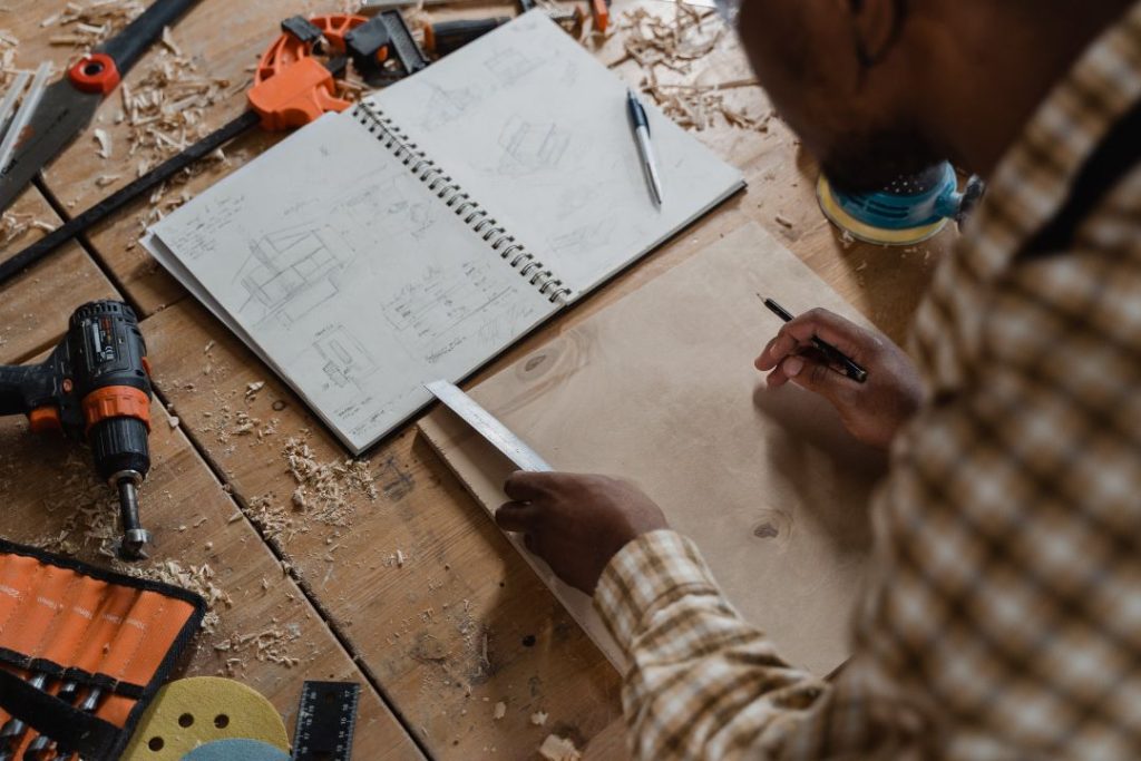 Man measuring a piece of wood while referencing rouch sketches in a book